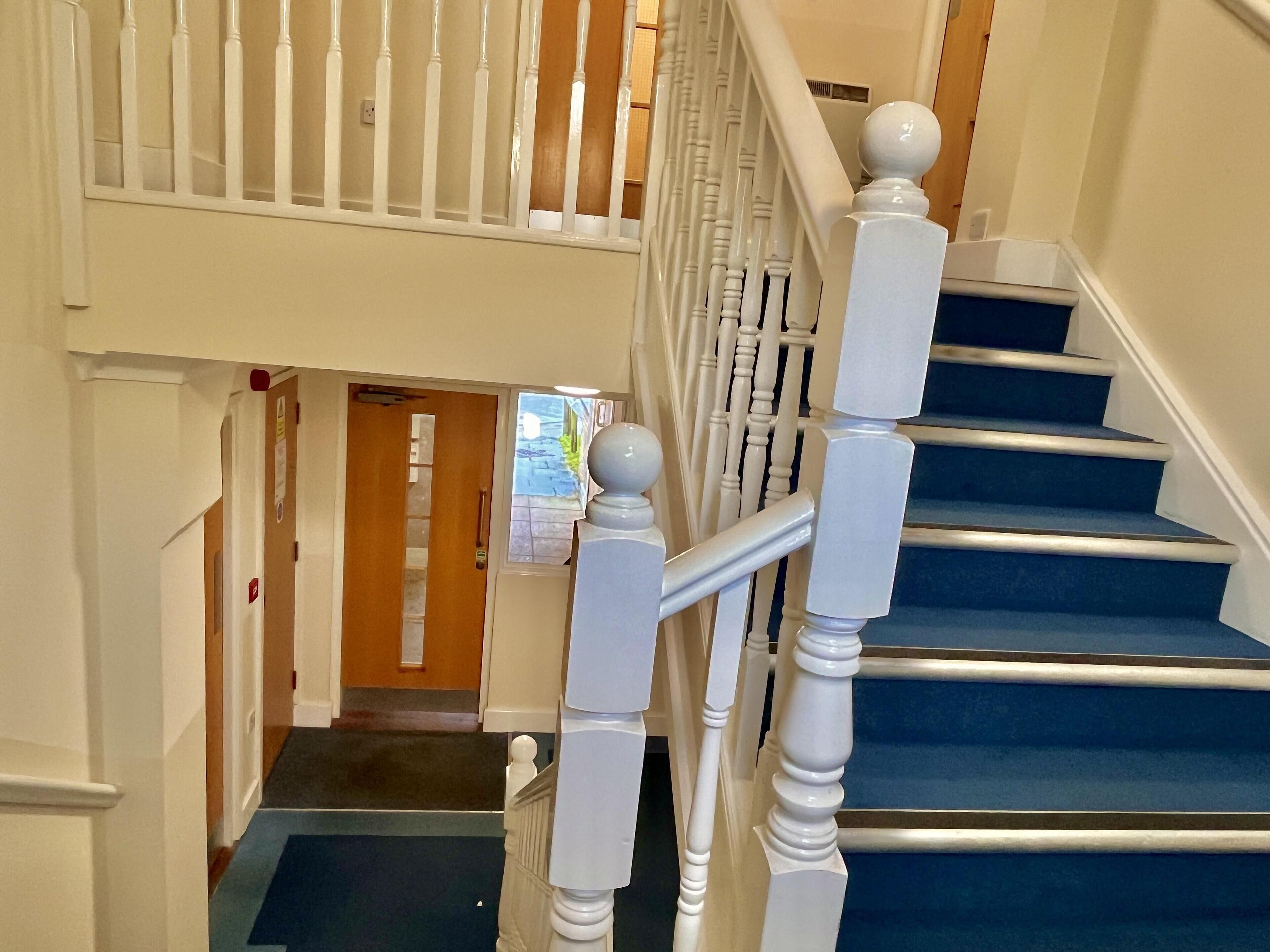 Un escalier avec des balustrades blanches et des marches recouvertes d’une moquette bleue, menant à un palier.