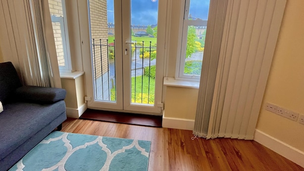 Une pièce avec un canapé aux couleurs sombres, un tapis à motifs et une vue, à travers un ensemble de portes vitrées, sur un balcon doté d’une balustrade, de plantes et de bâtiments en arrière-plan.