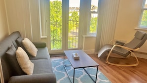 Un salon avec un canapé gris foncé, une petite table basse, un fauteuil à bascule et une vue sur un espace extérieur verdoyant, visible à travers de grandes fenêtres.