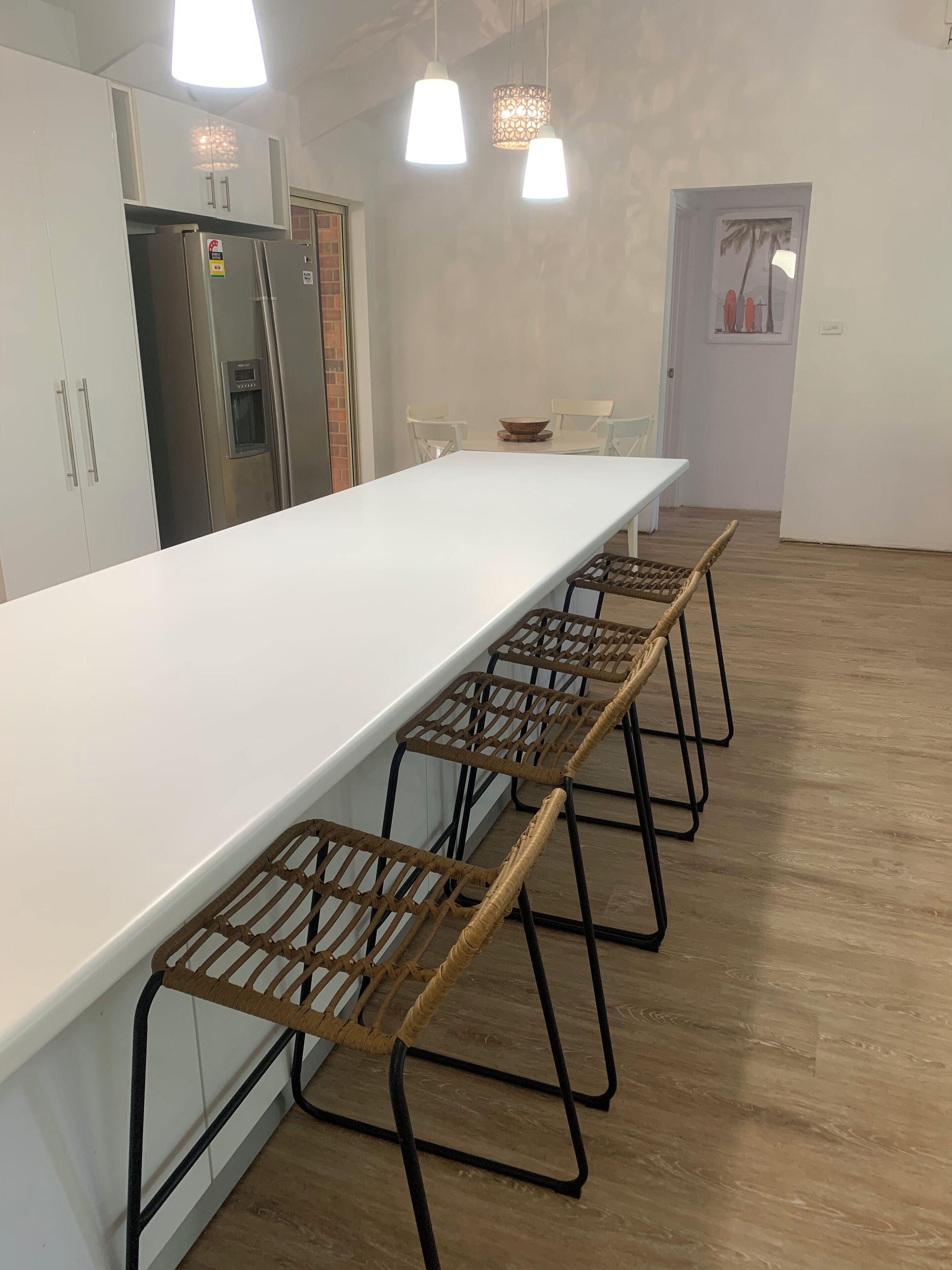 A modern kitchen with a long white countertop, stainless steel appliances, and a dining area with a long table and wicker chairs.