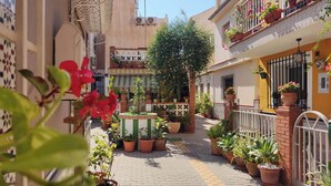 Un viuzza stretta con vasi di fiori e un albero al centro, affiancata da edifici con balconi e ringhiere.