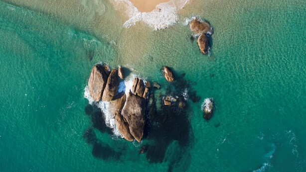 Eine Vogelperspektive zeigt einen Strand mit klarem türkisblauem Wasser, einem sandigen Ufer und mehreren großen Felsen, die aus dem Wasser ragen.