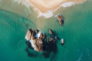 Eine Vogelperspektive zeigt einen Strand mit klarem türkisblauem Wasser, einem sandigen Ufer und mehreren großen Felsen, die aus dem Wasser ragen.