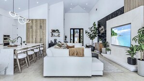 Een moderne woonkamer met een witte bank, een eethoek met een houten tafel en stoelen, een keuken met witte kasten, en een grote televisie aan de muur.