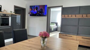 Una cocina moderna con horno, una mesa de comedor con un jarrón de flores, un sofá gris y un televisor montado en la pared.