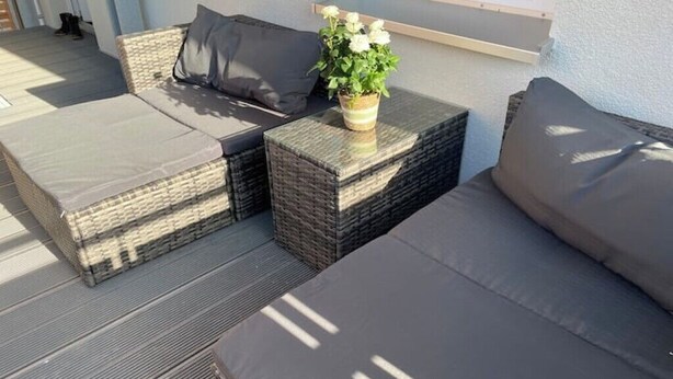 Un espacio al aire libre iluminado por el sol con sofás y cojines de mimbre, una pequeña mesa con una planta en maceta y una pared blanca al fondo.