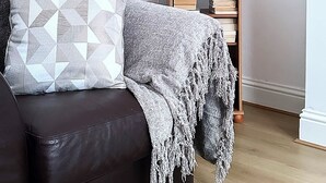 Un divano in pelle scura con cuscini decorati, una lampada bianca, un piccolo tavolino in legno con un vaso di pampa e una libreria.