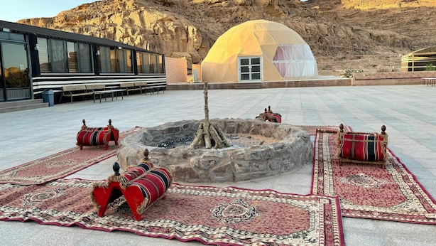 A desert landscape with a large rock formation in the background, a tent, and a fire pit with seating. - Jabal Rum camp (Wadi Rum)