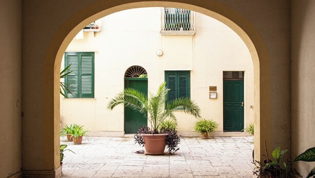 An arched courtyard with a paved floor, potted plants, and buildings with green shutters and doors.