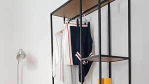 A room with a black metal shelving unit against a white wall, a grey chair with a floral cushion, and a floor lamp.