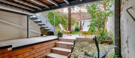 A covered outdoor area with a wooden staircase leading to a deck, surrounded by plants and a small garden.