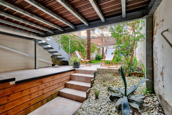 A covered outdoor area with a wooden staircase leading to a deck, surrounded by plants and a small garden.
