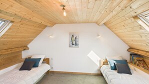 Une chambre confortable située sous les toits, avec deux lits simples, des poutres en bois au plafond, un tableau représentant des poissons accroché au mur et une bibliothèque remplie de livres.