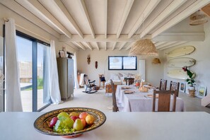 A dining area with a table set for two, a fruit bowl in the foreground, and surfboards on the wall.