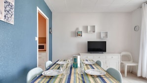 A dining area with a striped tablecloth, white chairs, and a television on a white cabinet. - Cozy apartment, loggia, beach 100 m - 4 guests (Saint-Hilaire-de-Riez)