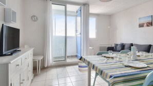 A modern, well-lit room with a dining table set for four, a television on a white cabinet, and a large window with curtains. - Cozy apartment, loggia, beach 100 m - 4 guests (Saint-Hilaire-de-Riez)