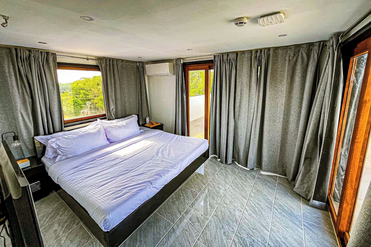 Une chambre à coucher avec un lit, des rideaux, un appareil de climatisation et une vue sur la verdure.