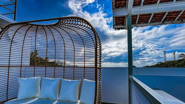 Ein moderner Außenbereich mit Sitzgelegenheiten, einer gebogenen Metallstruktur und weißen Kissen, mit Blick auf einen Pool und einen klaren Himmel.