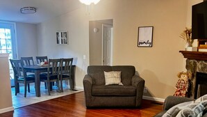 Un salon avec un canapé gris foncé, une table à manger en bois avec des chaises, un ventilateur de plafond et une cheminée agrémentée d’objets décoratifs.
