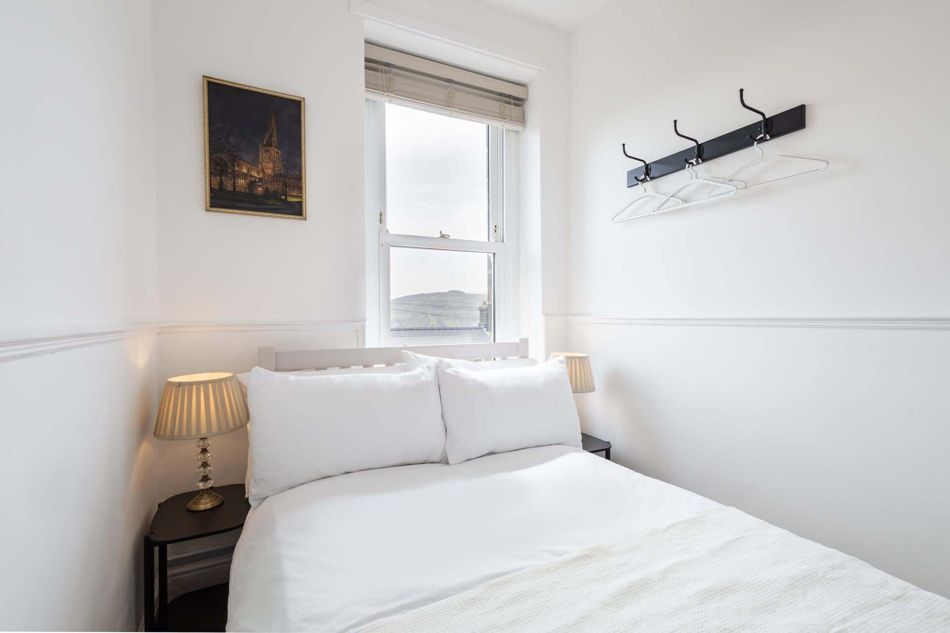A hotel room with a bed, two bedside lamps, a window with a view, a coat rack, and a framed picture on the wall.