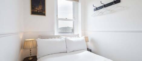 A hotel room with a bed, two bedside lamps, a window with a view, a coat rack, and a framed picture on the wall.