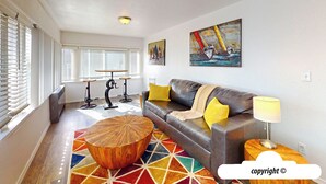 A modern living room with a leather sofa, a wooden coffee table, and a colorful rug. There are two paintings on the wall and a window with blinds. - Margarita's at Seaside Beach (Seaside)