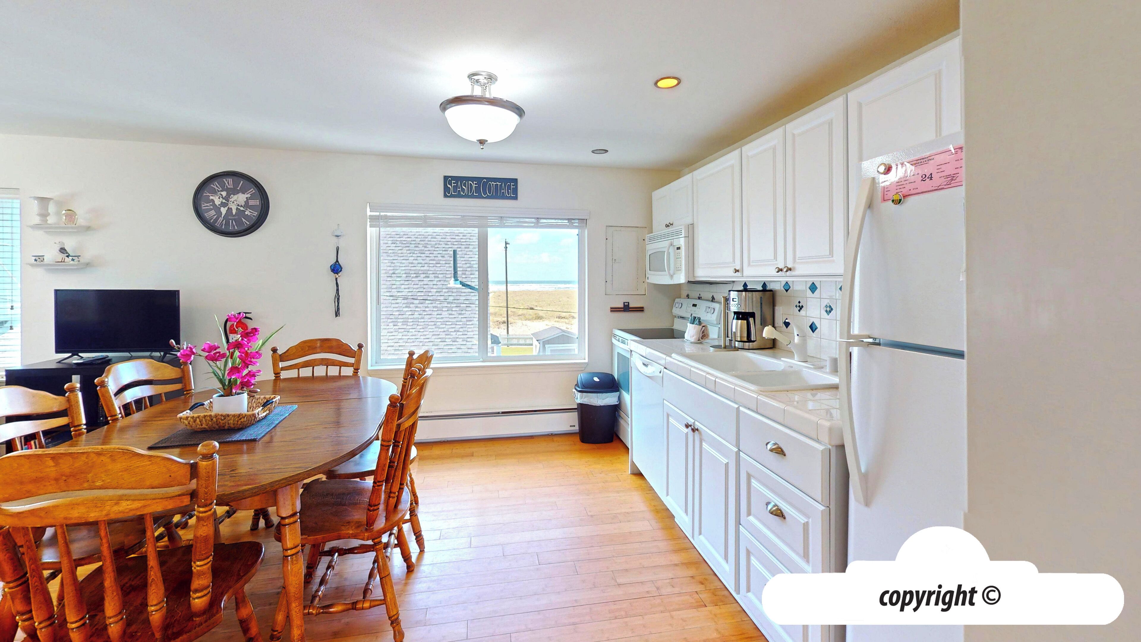 A compact kitchen with white cabinets, a dining table with chairs, a clock on the wall, and a view of the beach through the window.
