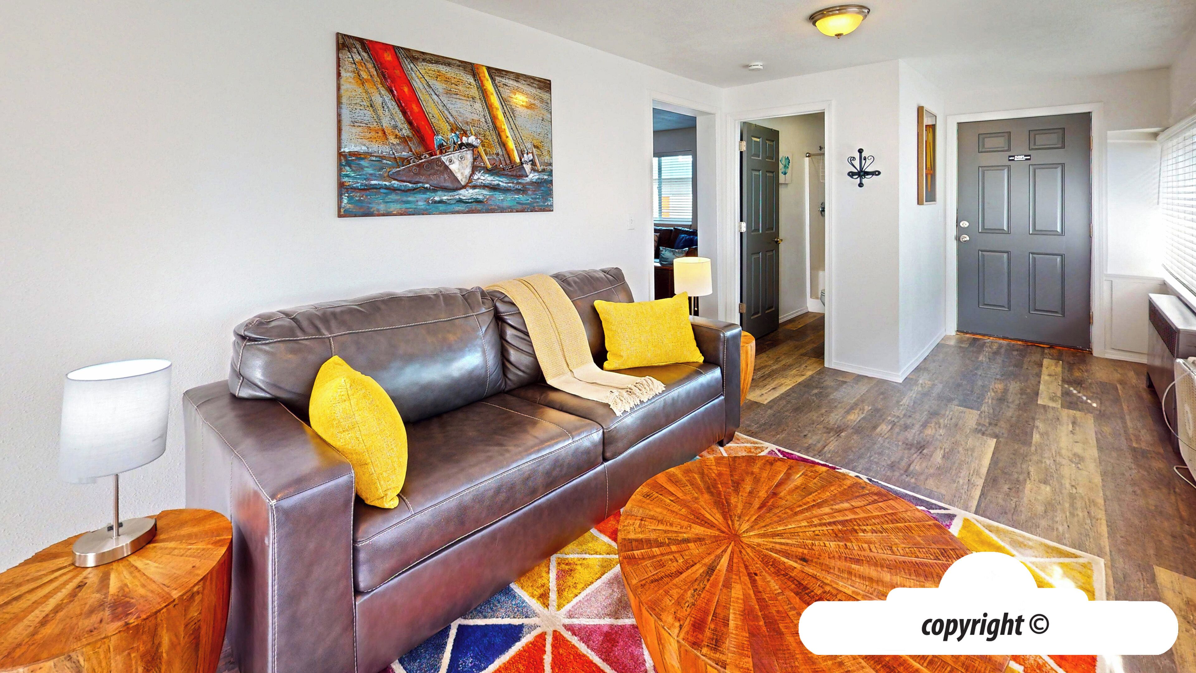 A modern living room with a brown leather sofa, a colorful rug, and a painting of a sailboat on the wall.