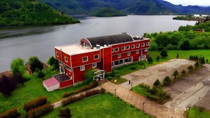 A red building with multiple windows and a flat roof, surrounded by greenery and a body of water. - ayvacık Hotel Restoran (Ayvacik)