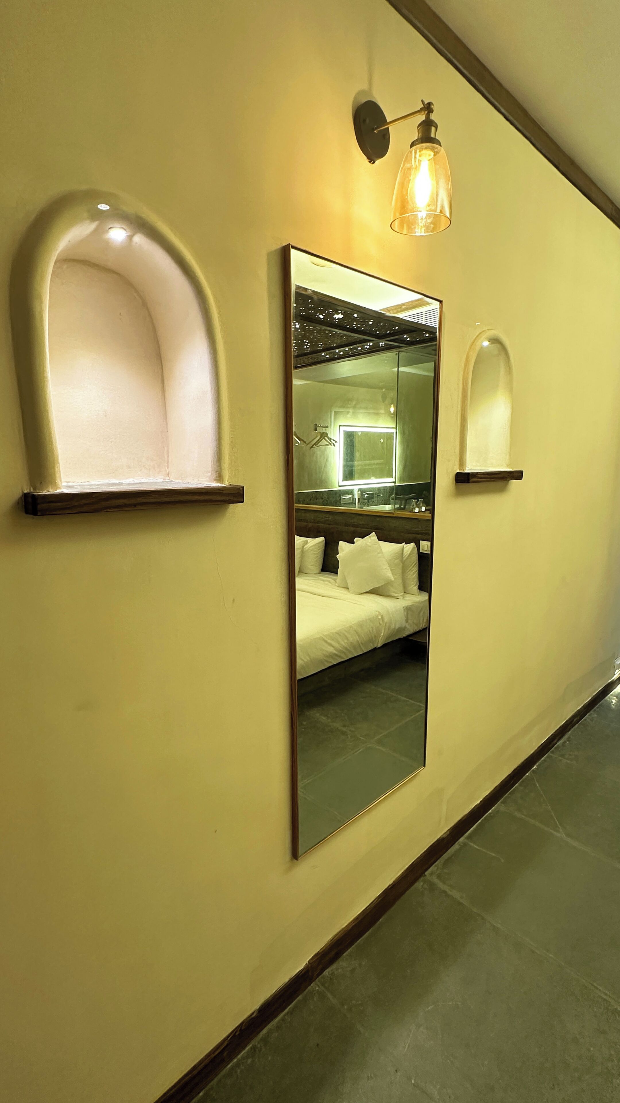 A hotel corridor with a large mirror reflecting a bed and a bathroom, and two wall-mounted sconces.