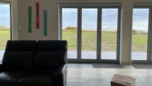 A modern living room with a black leather sofa, large sliding glass doors leading to a grassy outdoor area, and abstract wall art. - Relax at "Rocky Point" - a private, modern home w/cliff views of the Tasman Sea. (Cape Foulwind)