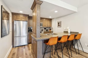 A kitchen with a stainless steel refrigerator, wooden cabinets, a granite countertop, and four orange bar stools.