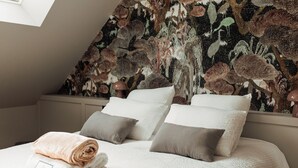 A neatly made bed with white and grey bedding, a rolled towel, and a framed picture on the bedside table. The room features a sloped ceiling and a wall with a detailed tapestry. - l'empreinte  (Verzenay)