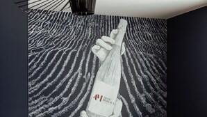 A modern living room with a dark grey sofa, two mustard-colored cushions, and a large wall mural depicting a bottle of champagne. - l'empreinte  (Verzenay)