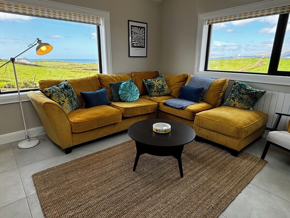 A living room with a mustard-colored sofa, a black coffee table, and a view of a green landscape through large windows.