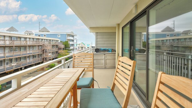 A balcony with wooden furniture, a view of a building with multiple windows, and a clear sky.