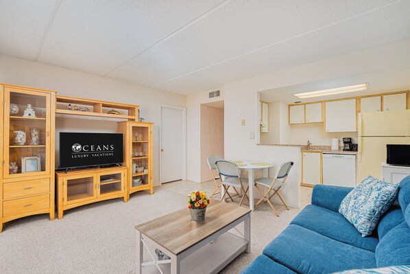A modern living room with a television, wooden cabinets, a dining area, and a kitchenette.