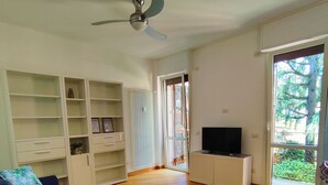 Uma sala de estar com sofá azul, estantes brancas, ventilador de teto, TV em um móvel e grandes janelas com vista para árvores.