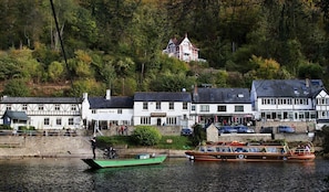 Una cittadina sulle rive del fiume, con edifici tradizionali, tra cui un pub bianco e una casa con tetto rosso.