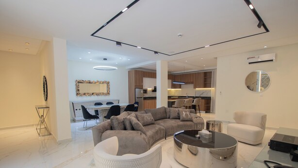 A modern living room with a sectional sofa, a round coffee table, and a dining area visible in the background. - Mini Palace Luxury Suites (Abuja)