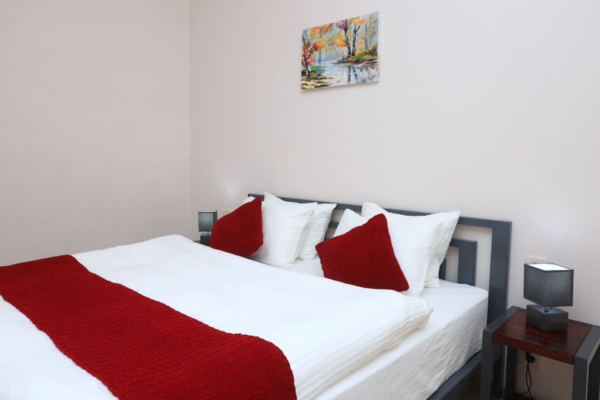 A neatly made bed with white and red bedding, a painting on the wall, and a bedside table with a lamp.