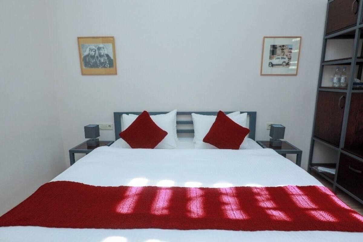 A neatly made bed with a white and red bedspread, two red pillows, and a red throw blanket. There are two bedside tables with lamps, and a bookshelf to the right.