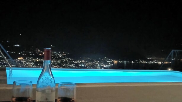 Une table avec une bouteille et des verres, placée au bord d’une piscine, avec une vue sur la ville illuminée la nuit.