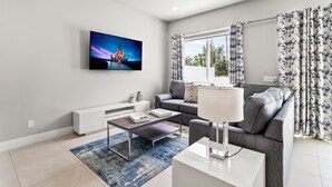Een moderne woonkamer met een flatscreen televisie, een grijze bank, een witte salontafel en behang met een patroon.