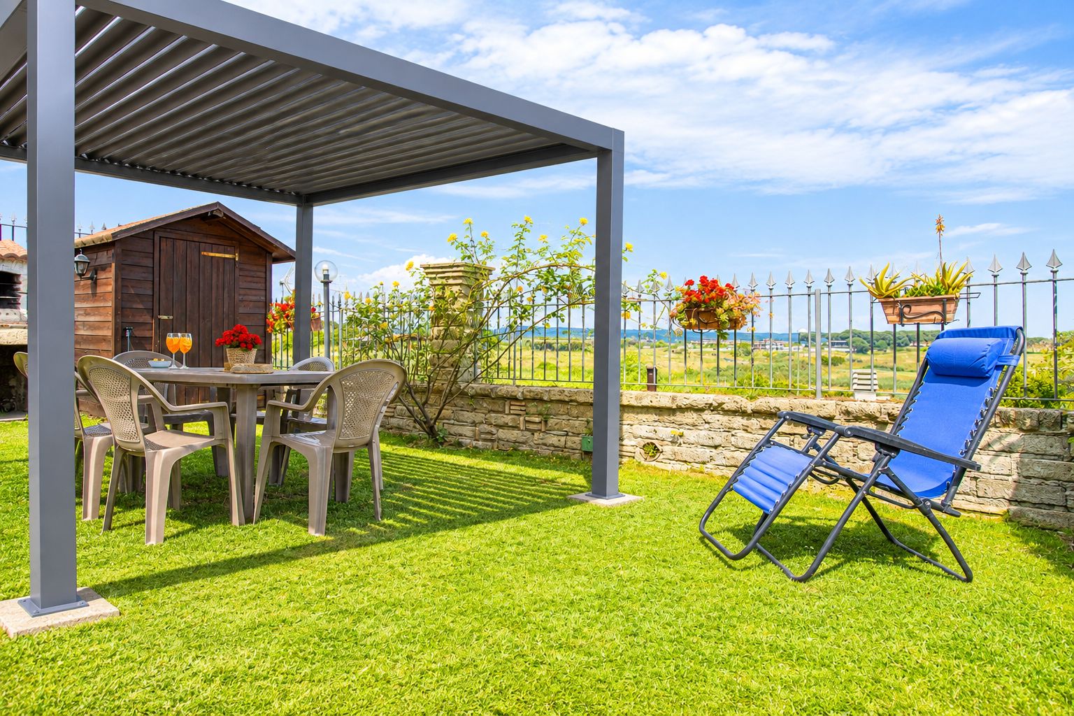A covered outdoor seating area with a table and chairs, a blue lounge chair, and a stone wall with a view of the countryside.