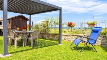A covered outdoor seating area with a table and chairs, a blue lounge chair, and a stone wall with a view of the countryside.
