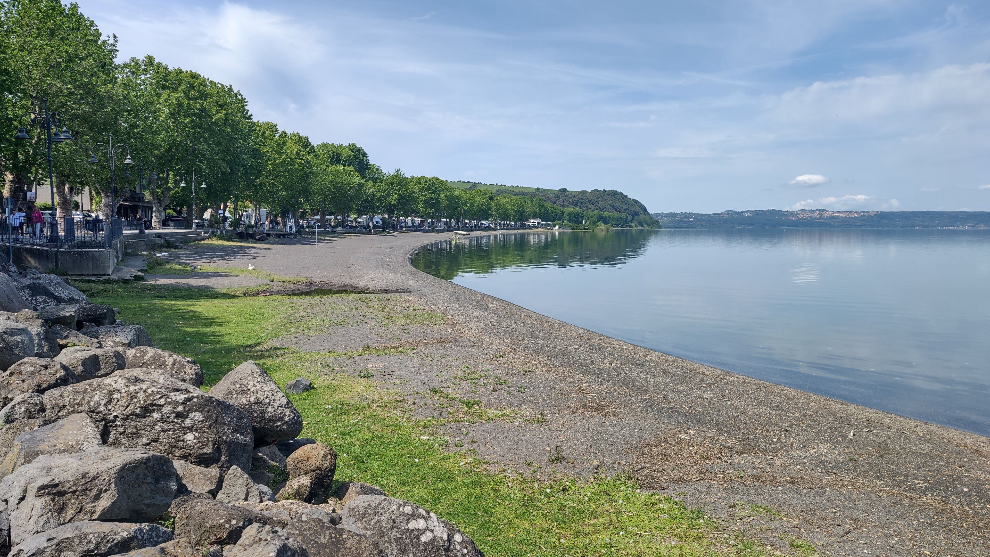 A lakeside park with a paved walkway, picnic areas, and a row of trees.