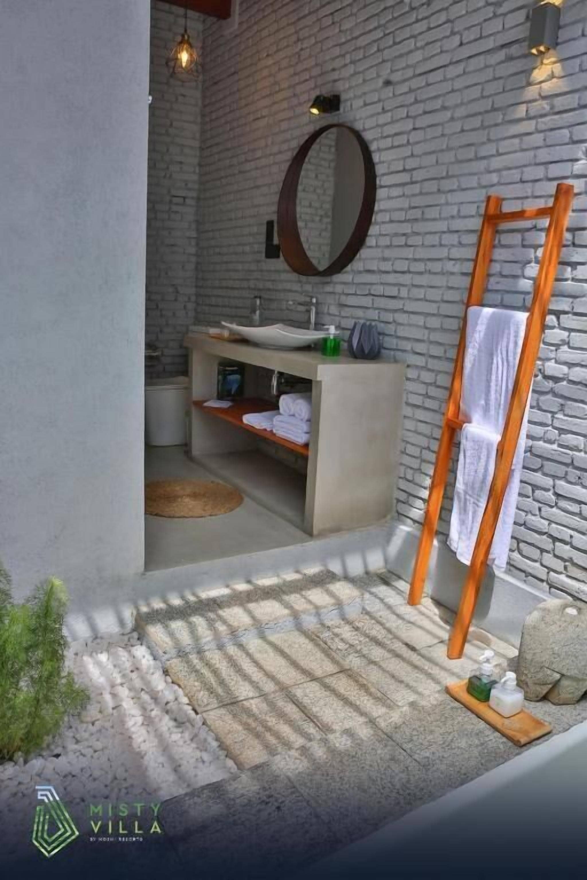 Un baño moderno con pared de piedra, tocador de madera, espejo redondo y perchero de madera.