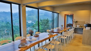 Un espace repas avec une longue table en bois, des chaises blanches et une vue sur un paysage luxuriant, visible à travers de grandes fenêtres.