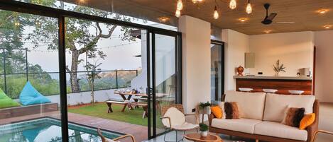 A modern living room with a glass wall, a pool, and outdoor furniture.